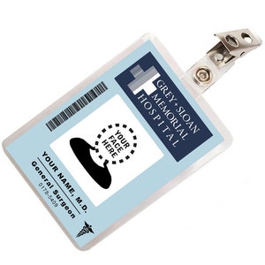 May include: A blue and white plastic ID badge holder with a clip. The badge holder has the text "GREY+SLOAN MEMORIAL HOSPITAL" printed on it. The badge holder has a space for a photo with the text "YOUR FACE HERE" printed around it.