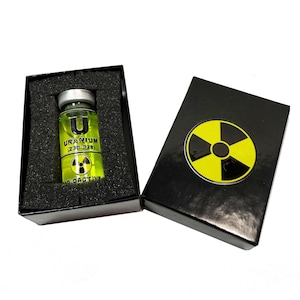 Puede incluir: Un pequeño vial de vidrio con un líquido amarillo en su interior. El vial tiene una etiqueta negra que dice "URANIUM (230-028)" y un símbolo de radiactividad. El vial está en una caja negra con un símbolo de radiactividad en la tapa.