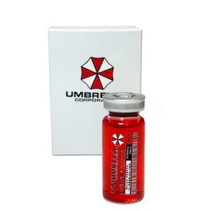 Op de afbeelding: Een klein glazen flesje gevuld met een rode vloeistof. Het flesje heeft een zilveren dop en een etiket met de tekst "UMBRELLA CORPORATION T-VIRUS" met een barcode en het nummer "01".