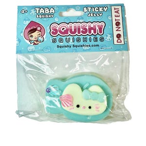 May include: A packaged squishy toy in a clear plastic wrapper. The squishy is shaped like a light green heart with a white face, pink seashell, and a blue bow. The packaging has the words "Squishy Squishies" and "Sticky Jelly".