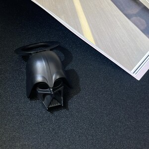 Puede incluir: Un abridor de botellas negro con forma de casco de Darth Vader, con un asa circular. El abridor est&aacute; sobre una superficie oscura, con una revista en el fondo.