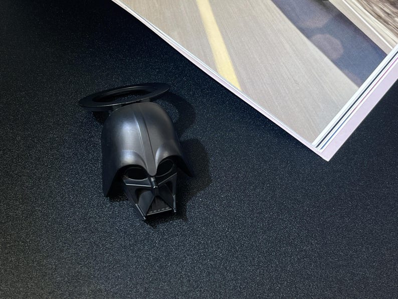 Puede incluir: Un abridor de botellas negro con forma de casco de Darth Vader, con un asa circular. El abridor est&aacute; sobre una superficie oscura, con una revista en el fondo.