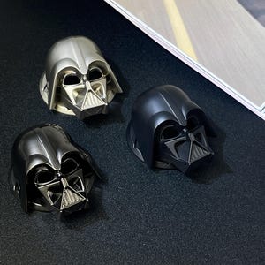 Puede incluir: Tres tapones de botella con forma de casco de Darth Vader en plata, negro y gris plomo. Los tapones presentan el ic&oacute;nico dise&ntilde;o del casco de la franquicia Star Wars. Los tapones est&aacute;n sobre una superficie oscura.
