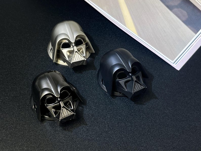 Puede incluir: Tres tapones de botella con forma de casco de Darth Vader en plata, negro y gris plomo. Los tapones presentan el ic&oacute;nico dise&ntilde;o del casco de la franquicia Star Wars. Los tapones est&aacute;n sobre una superficie oscura.