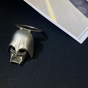 Puede incluir: Un abrebotellas plateado con forma de casco de Darth Vader, con un asa circular. El abrebotellas est&aacute; sobre una superficie negra, con una imagen enmarcada en el fondo.