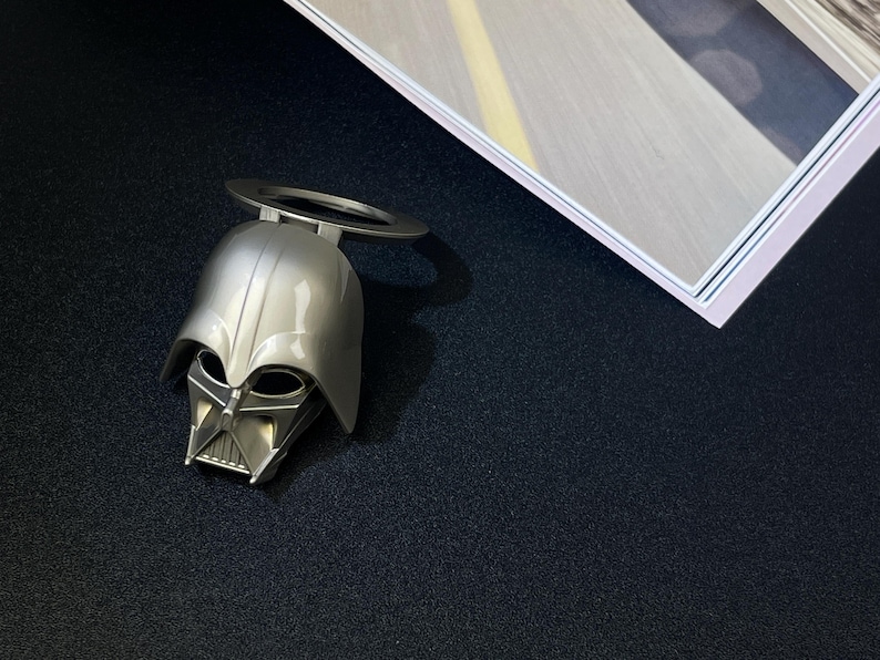 Puede incluir: Un abrebotellas plateado con forma de casco de Darth Vader, con un asa circular. El abrebotellas est&aacute; sobre una superficie negra, con una imagen enmarcada en el fondo.