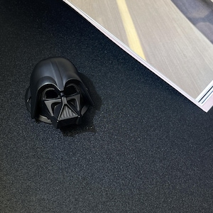 Puede incluir: Una figura de casco negro de Darth Vader. El casco es una r&eacute;plica detallada del ic&oacute;nico personaje de Star Wars, con un acabado brillante. La figura est&aacute; colocada sobre una superficie oscura y texturizada, con una porci&oacute;n de una revista visible en el fondo.