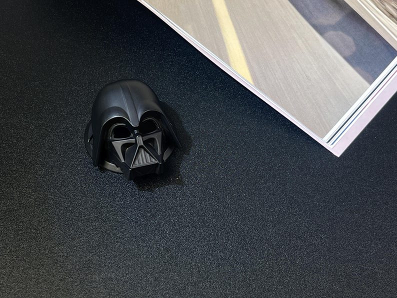 Puede incluir: Una figura de casco negro de Darth Vader. El casco es una r&eacute;plica detallada del ic&oacute;nico personaje de Star Wars, con un acabado brillante. La figura est&aacute; colocada sobre una superficie oscura y texturizada, con una porci&oacute;n de una revista visible en el fondo.