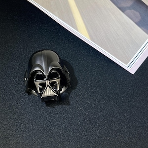 Puede incluir: Una figura met&aacute;lica del casco de Darth Vader, con un dise&ntilde;o detallado de la m&aacute;scara del personaje ic&oacute;nico. El casco es principalmente negro con detalles plateados en la cara, sobre un fondo oscuro.