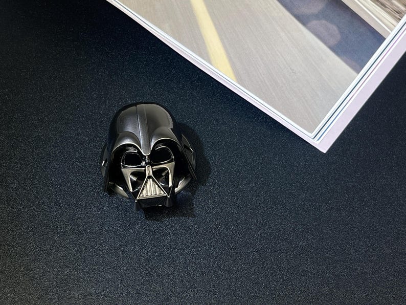 Puede incluir: Una figura met&aacute;lica del casco de Darth Vader, con un dise&ntilde;o detallado de la m&aacute;scara del personaje ic&oacute;nico. El casco es principalmente negro con detalles plateados en la cara, sobre un fondo oscuro.