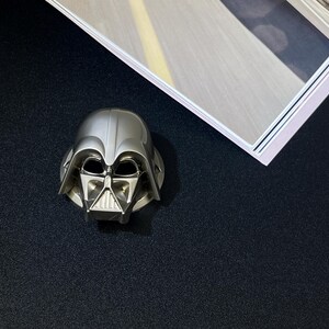 Puede incluir: Figura de casco de Darth Vader plateada. El casco presenta detalles intrincados de la m&aacute;scara y los rasgos faciales. La figura se coloca sobre una superficie oscura, con una imagen enmarcada visible en la esquina superior derecha.