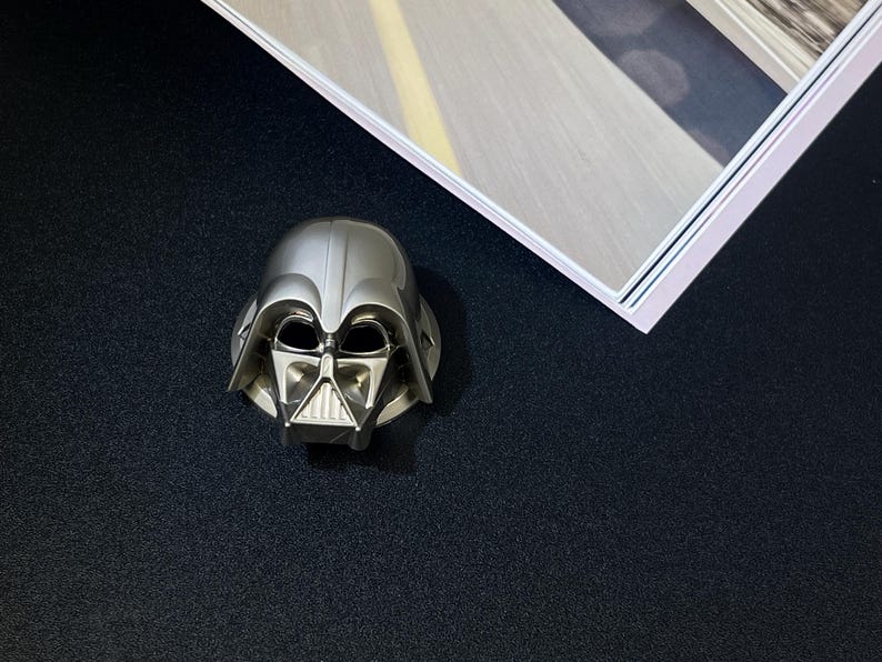 Puede incluir: Figura de casco de Darth Vader plateada. El casco presenta detalles intrincados de la m&aacute;scara y los rasgos faciales. La figura se coloca sobre una superficie oscura, con una imagen enmarcada visible en la esquina superior derecha.