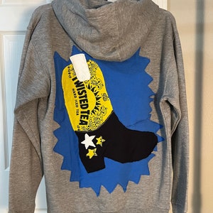 Peut inclure: Sweat-shirt à capuche gris avec un motif bleu et jaune d'une botte avec le texte "Twisted Tea Hard Iced Tea" au dos.