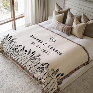 May include: Cream-colored woven throw blanket with black floral design along the left edge. The blanket features the names "Amanda & Connor", a heart symbol, and the date "06-23-2025" in black text. The blanket has a fringed edge.