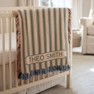 May include: A beige and gray striped baby blanket with the name "THEO SMITH" in a rectangular label is draped over a white crib. The blanket has a fringed edge with a mix of colors. The nursery has a white crib, a white chair, and a window.