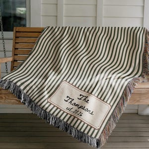 May include: A woven throw blanket featuring a pattern of vertical dark green and cream stripes. A rectangular patch in the lower corner displays the text The Thompsons est. 2026 in black script. The blanket has frayed edges along the bottom border.