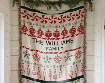 May include: A cream-colored Christmas throw blanket with red and green designs. The blanket features candy canes, ornaments, snowflakes, and Christmas trees. The text "THE WILLIAMS FAMILY" is printed in black.