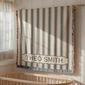 May include: A beige and gray striped blanket with the name "THEO SMITH" displayed in a gray banner. The blanket is hanging on a wooden rod, with a wooden crib visible below. The blanket has fringed edges.