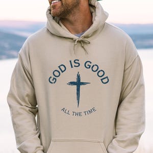 May include: Sand-colored hoodie with a front pocket and drawstring hood. The front features the text "GOD IS GOOD ALL THE TIME" in navy blue, with a cross graphic. The hoodie is a casual, comfortable garment.
