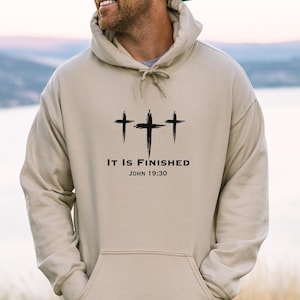 May include: A sand-colored hoodie with a drawstring hood and a kangaroo pocket. The front features three black crosses above the text "IT IS FINISHED" and "JOHN 19:30". The hoodie is worn by a person.