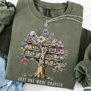 Puede incluir: Sudadera verde militar con un dise&ntilde;o de &aacute;rbol floral y estanter&iacute;as. El texto "JUST ONE MORE CHAPTER" est&aacute; impreso debajo del &aacute;rbol. Un collar plateado est&aacute; sobre la sudadera. La sudadera est&aacute; doblada junto a unos vaqueros azules.