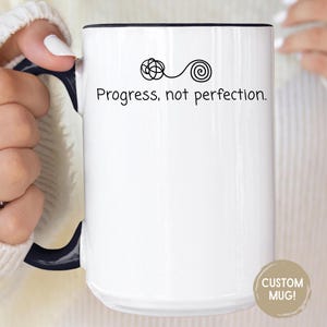 May include: White ceramic mug with a black rim and handle. The mug features the text "Progress, not perfection." in a black, minimalist font, along with a small graphic. The mug is held by a person wearing a white sweater.