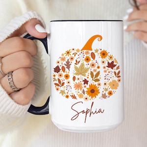 May include: White ceramic mug with a black interior and handle. The mug features a pumpkin design made of fall-colored flowers and leaves. The name "Sophia" is written in a script font below the pumpkin.