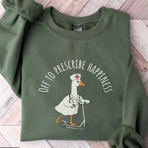 May include: Military green sweatshirt with the text "OFF TO PRESCRIBE HAPPINESS" arched above a cartoon goose wearing a nurse's hat and coat, riding a scooter. The sweatshirt has long sleeves and a crew neck.