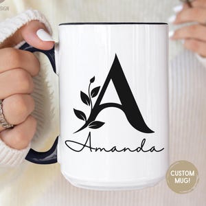 May include: White ceramic mug with a black interior and handle. The mug features a large black letter "A" with a floral design and the name "Amanda" in script. A small, round sticker on the mug reads "CUSTOM MUG!"