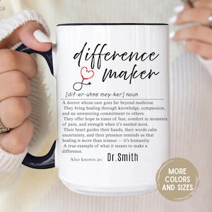 May include: White ceramic mug with a black handle and rim, featuring the words "difference maker" in black script, a red heart and stethoscope graphic, and a definition. The mug also includes the name "Dr. Smith".