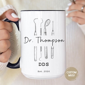 May include: White ceramic mug with a black rim and handle, featuring dental tools illustrations. The mug is personalized with the name "Dr. Thompson", the initials "DDS", and the year "Est. 2026". The mug has a "Custom Mug!" sticker.