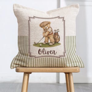 May include: A square throw pillow with a textured beige fabric featuring a central illustration of a teddy bear wearing a cap and playing golf. The bear stands on a green patch next to a golf bag. The name Oliver is printed below in brown.