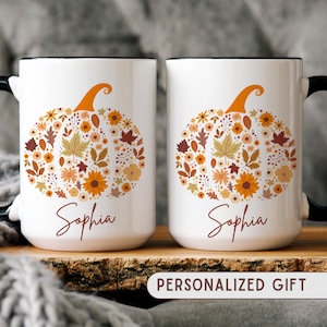 May include: Two white ceramic mugs with black handles and rims. Each mug features a floral pumpkin design in orange, yellow, and brown, with the name "Sophia" below. The mugs are labeled "PERSONALIZED GIFT".