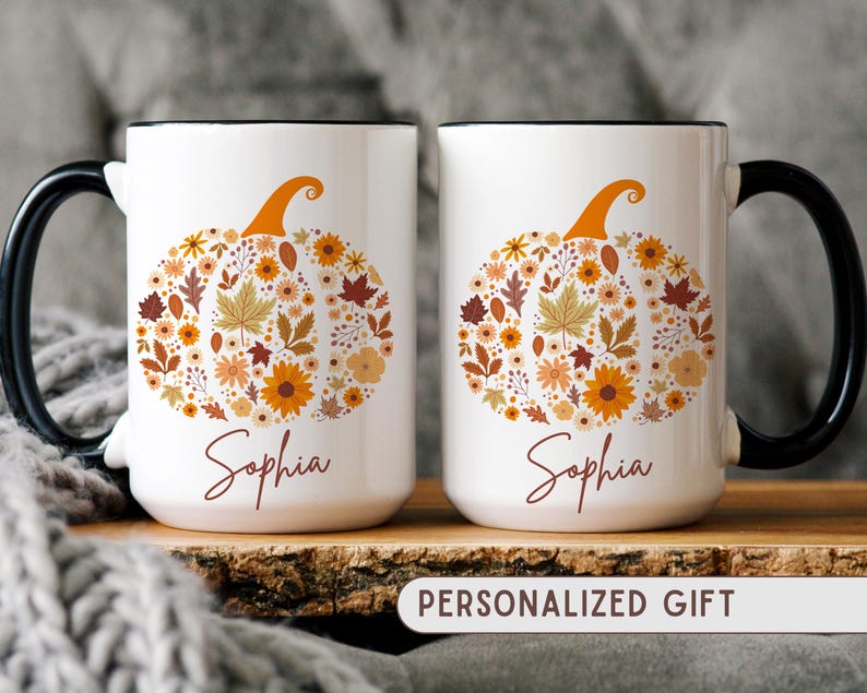 May include: Two white ceramic mugs with black handles and rims. Each mug features a floral pumpkin design in orange, yellow, and brown, with the name "Sophia" below. The mugs are labeled "PERSONALIZED GIFT".