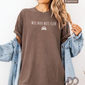 May include: Espresso-colored t-shirt with rolled sleeves. The shirt features the text "Wee Woo Wife Club" above a car graphic. The model is wearing a denim jacket over the shirt. The Dizquiel Design logo is visible in the bottom right corner.
