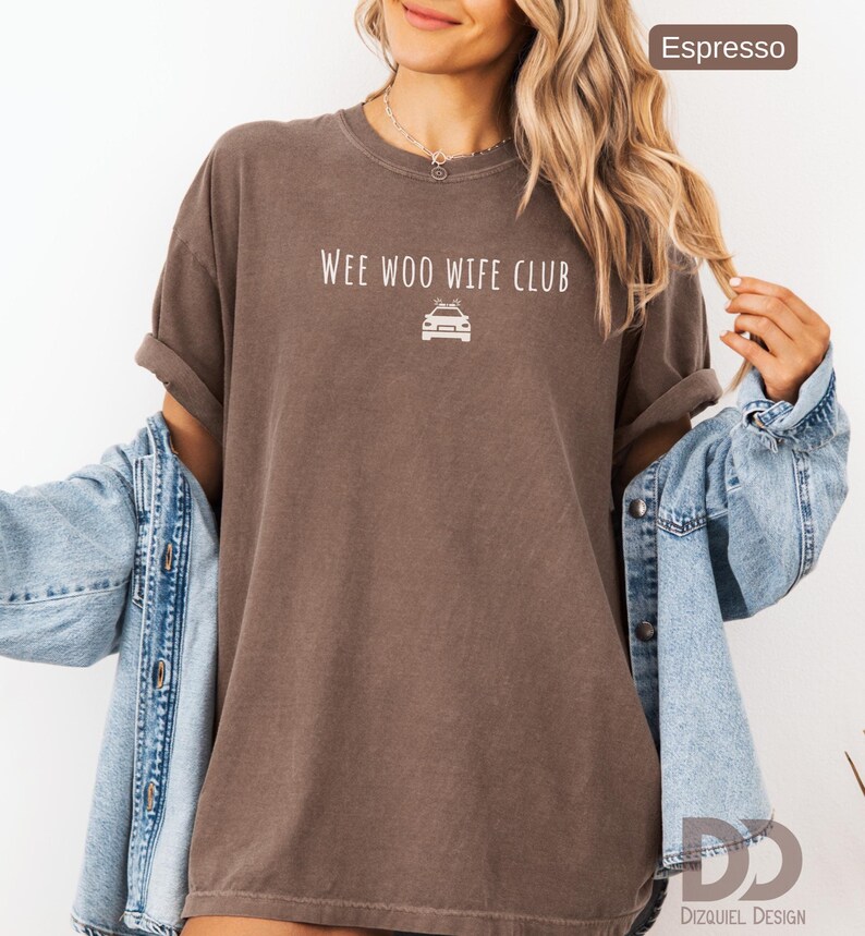 May include: Espresso-colored t-shirt with rolled sleeves. The shirt features the text "Wee Woo Wife Club" above a car graphic. The model is wearing a denim jacket over the shirt. The Dizquiel Design logo is visible in the bottom right corner.