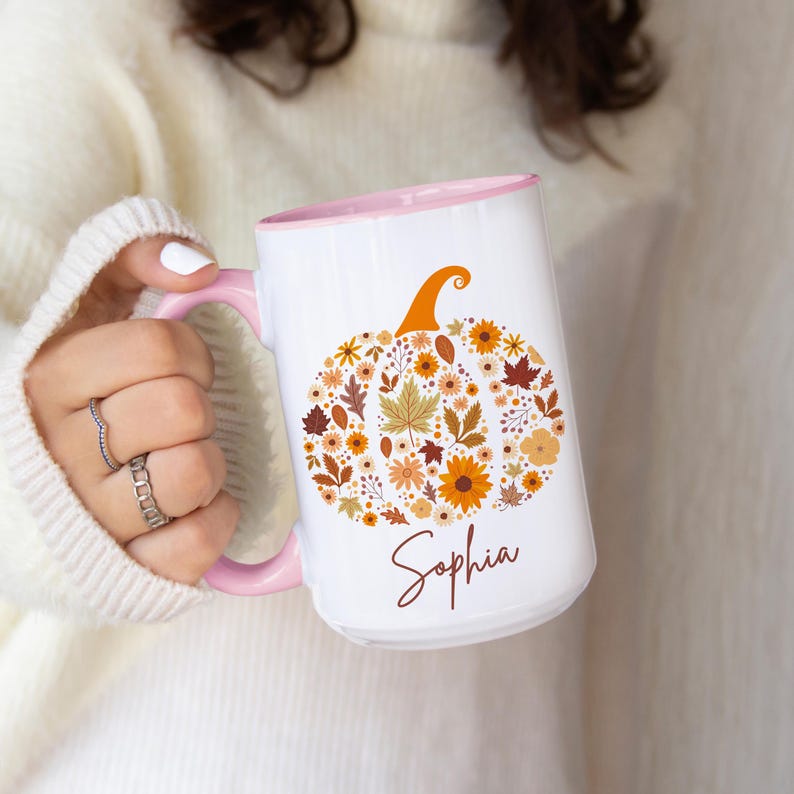 May include: White ceramic mug with a pink handle and interior. The mug features a pumpkin design made of fall leaves and flowers in shades of orange, brown, and yellow. The name "Sophia" is written in a script font.