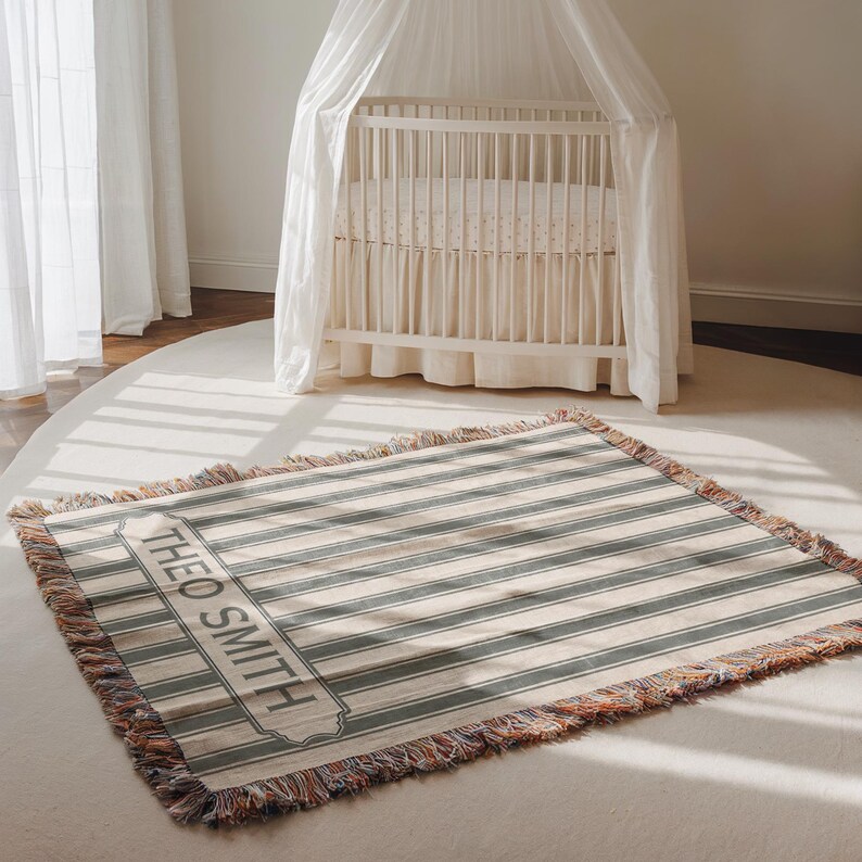 May include: A rectangular throw blanket with horizontal stripes in muted blue-green and cream. The blanket has a fringed edge and a rectangular label with the name "THEO SMITH" printed on it. A white crib with a canopy is in the background.
