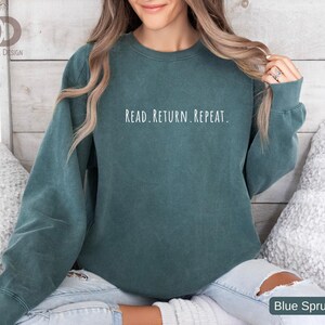 May include: A teal-colored sweatshirt with the text "READ. RETURN. REPEAT." in white lettering. The sweatshirt is a solid color and has long sleeves. The bottom right corner of the image has the text "Blue Spruce".