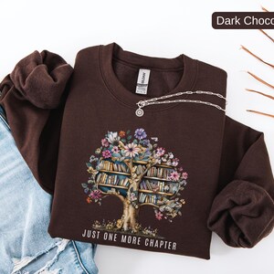 Puede incluir: Sudadera de color chocolate oscuro con un dise&ntilde;o de &aacute;rbol de libros y flores. El gr&aacute;fico incluye un tronco de &aacute;rbol con estanter&iacute;as y flores coloridas. El texto "JUST ONE MORE CHAPTER" est&aacute; impreso en la parte inferior. Un collar plateado est&aacute; sobre la sudadera.