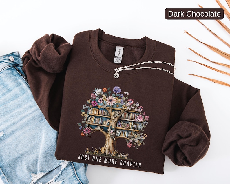 Puede incluir: Sudadera de color chocolate oscuro con un dise&ntilde;o de &aacute;rbol de libros y flores. El gr&aacute;fico incluye un tronco de &aacute;rbol con estanter&iacute;as y flores coloridas. El texto "JUST ONE MORE CHAPTER" est&aacute; impreso en la parte inferior. Un collar plateado est&aacute; sobre la sudadera.