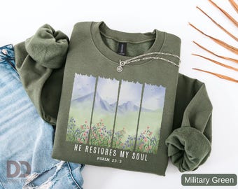 May include: Military green sweatshirt with a landscape graphic featuring mountains, flowers, and the text "HE RESTORES MY SOUL" and "PSALM 23:3". A silver necklace is draped across the sweatshirt. The sweatshirt is paired with blue jeans.