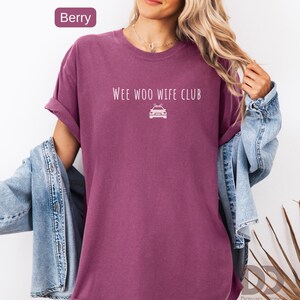 May include: Berry-colored t-shirt with rolled sleeves. The shirt features the text "WEE WOO WIFE CLUB" above a white graphic of a police car. A denim jacket is draped over the shoulders.