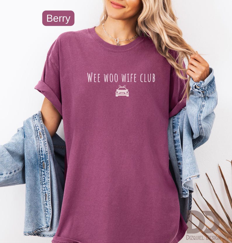 May include: Berry-colored t-shirt with rolled sleeves. The shirt features the text "WEE WOO WIFE CLUB" above a white graphic of a police car. A denim jacket is draped over the shoulders.
