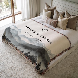 May include: A cream-colored throw blanket with a mountain landscape design in shades of gray and black. The blanket features the names "AMANDA & CONNOR", a heart symbol, and the date "03-06-2025". The blanket has a fringed edge and is displayed on a bed.