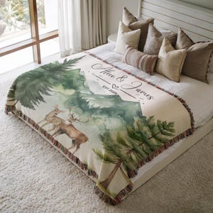 May include: A cream-colored throw blanket with a watercolor design of a mountain scene, trees, and deer. The blanket has a fringed edge and the text "Alice & James EST. 2026".