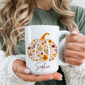 May include: White ceramic mug featuring a pumpkin design made of fall leaves and flowers in shades of orange, brown, and yellow. The name "Sophia" is written in a cursive font below the pumpkin. The mug has a large handle.