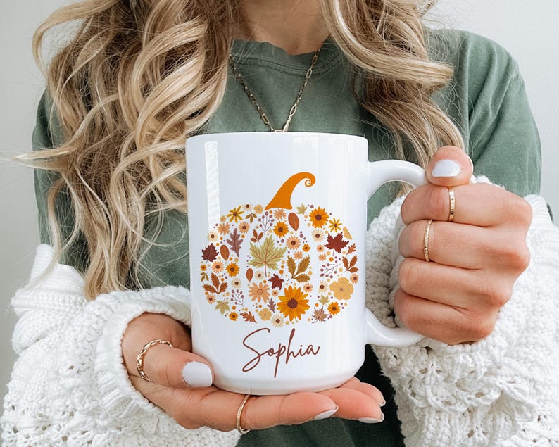 May include: White ceramic mug featuring a pumpkin design made of fall leaves and flowers in shades of orange, brown, and yellow. The name "Sophia" is written in a cursive font below the pumpkin. The mug has a large handle.
