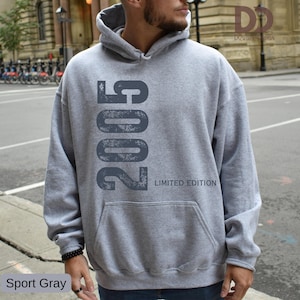 Pu&ograve; includere: Una felpa con cappuccio grigio chiaro con tasca frontale. La felpa presenta il numero "2005" stampato verticalmente in un carattere grigio scuro invecchiato, con "LIMITED EDITION" sotto. Il testo "Sport Gray" &egrave; in basso a sinistra.