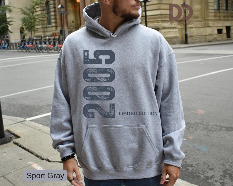 Pu&ograve; includere: Una felpa con cappuccio grigio chiaro con tasca frontale. La felpa presenta il numero "2005" stampato verticalmente in un carattere grigio scuro invecchiato, con "LIMITED EDITION" sotto. Il testo "Sport Gray" &egrave; in basso a sinistra.